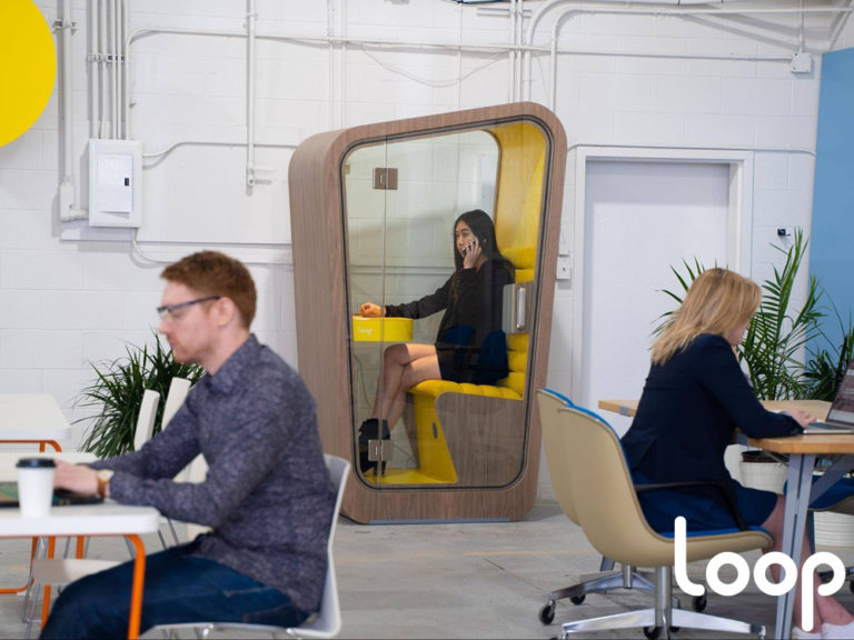 Employees working in an open office while one worker utilizes a Loop Phone Booth to make a private phone call