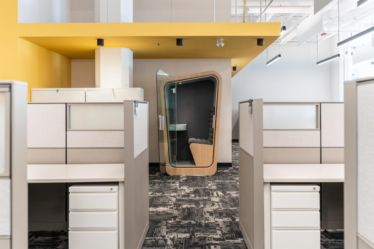 A custom loop solo phone booth sits inside an empty standard office with cubicles surronding it and yellow trim on the walls