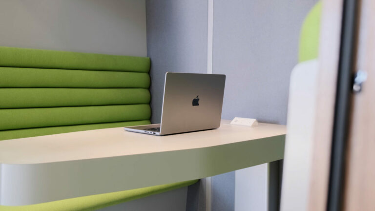A macbook pro is sitting on the desk of a loop cube meeting pod with green cloth seats