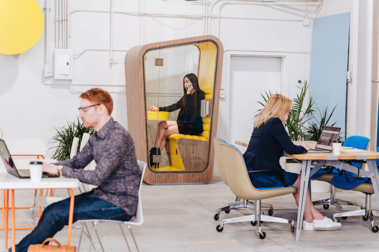 a woman takes a phone call in privacy using loop solo phone booth at her office with co workers working at desks around her